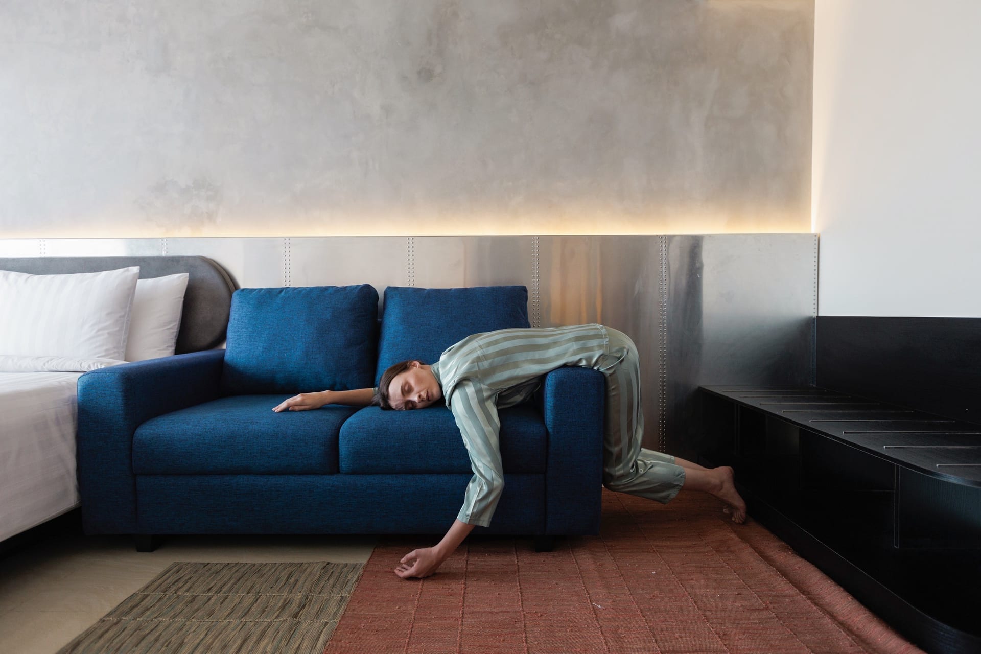 Guest in striped pyjamas asleep face-down across the blue sofa beside the bed