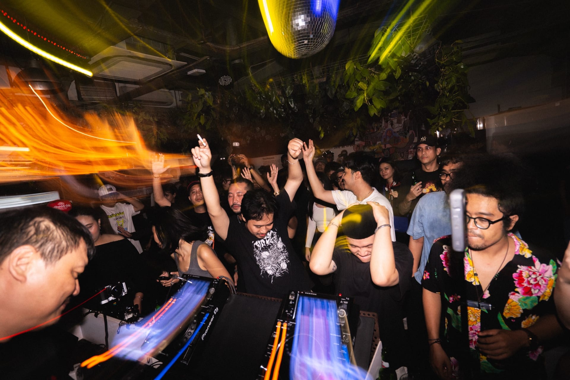 Dancers with arms raised behind the CDJ decks under a disco ball at 18+ Music Bar