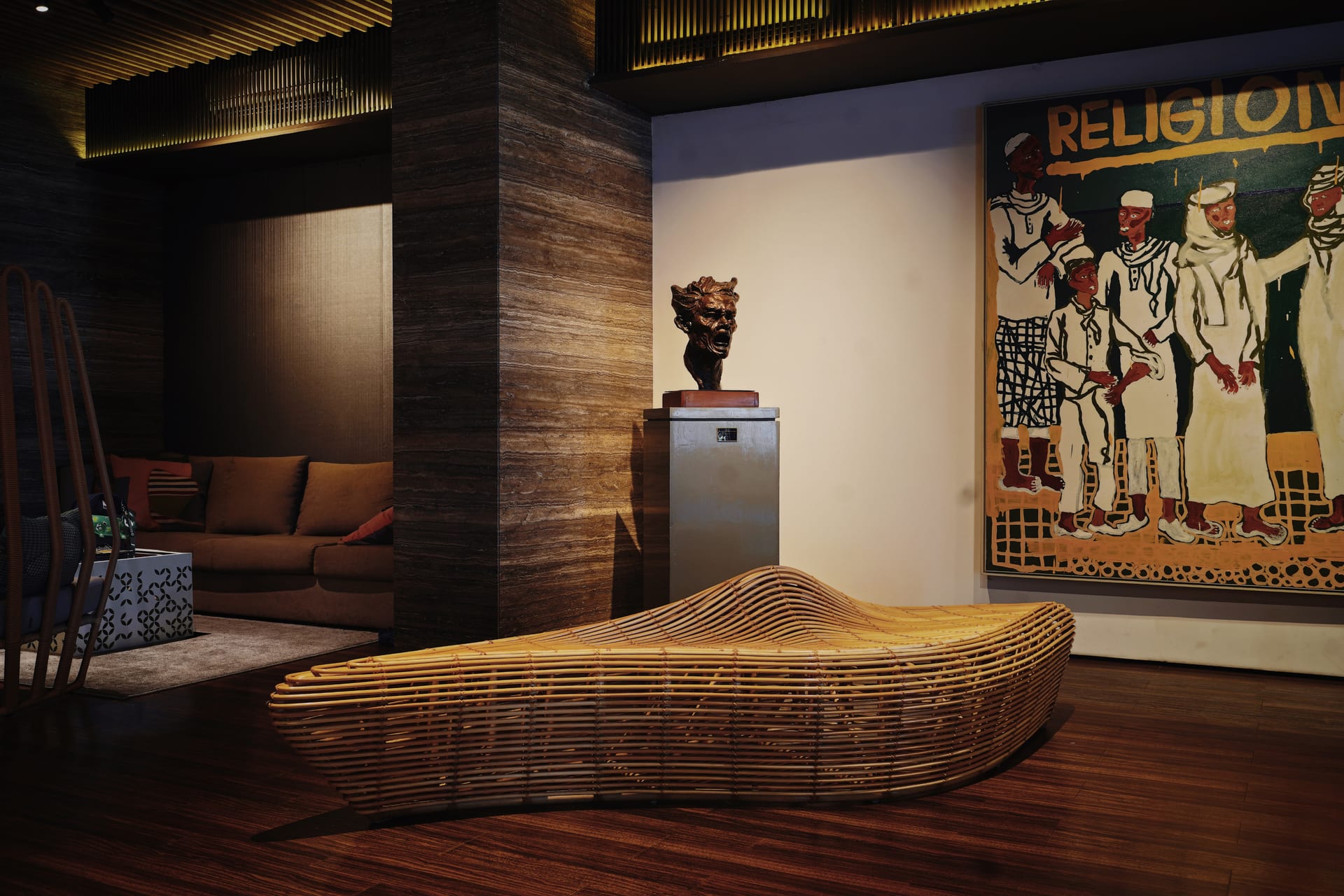 Sculptural rattan bench and a bronze head bust beside a large painting in the PULANG ke UTTARA lobby