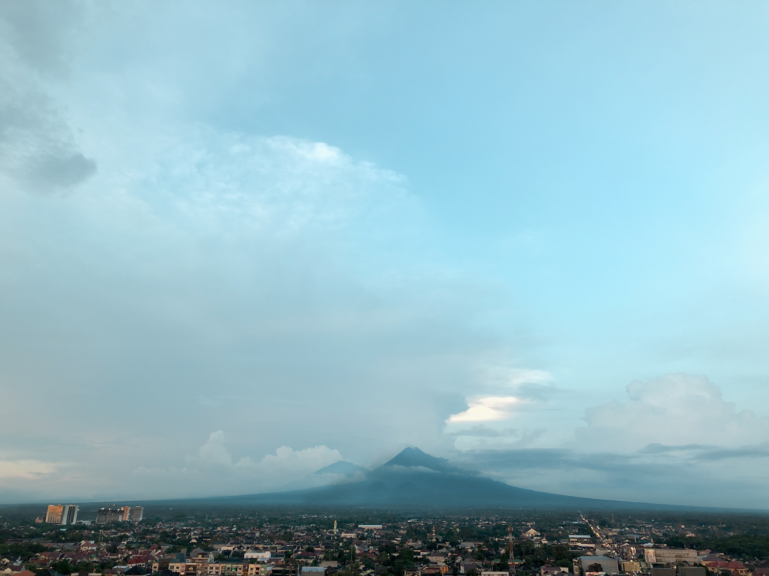 Mount Merapi rising over the rooftops of Jogja at dusk, seen from a high floor of UTTARA The Icon